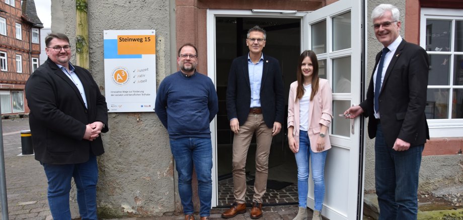 Vier Männer und eine Frau stehen vor einem Gebäude mit offener Tür. Am Gebäude hängt ein Schild, auf dem das Projekt A³ abgebildet ist.