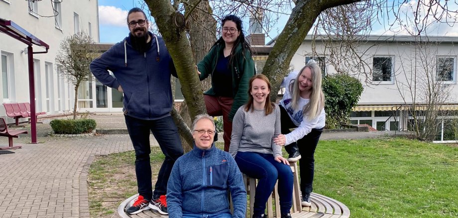 Gruppenfoto von fünf Personen. Ein Mann sitzt auf einer Holzbank, die einen Baum umfasst. Drei Frauen und ein Mann stehen auf der Sitzbank und lachen in die Kamera.