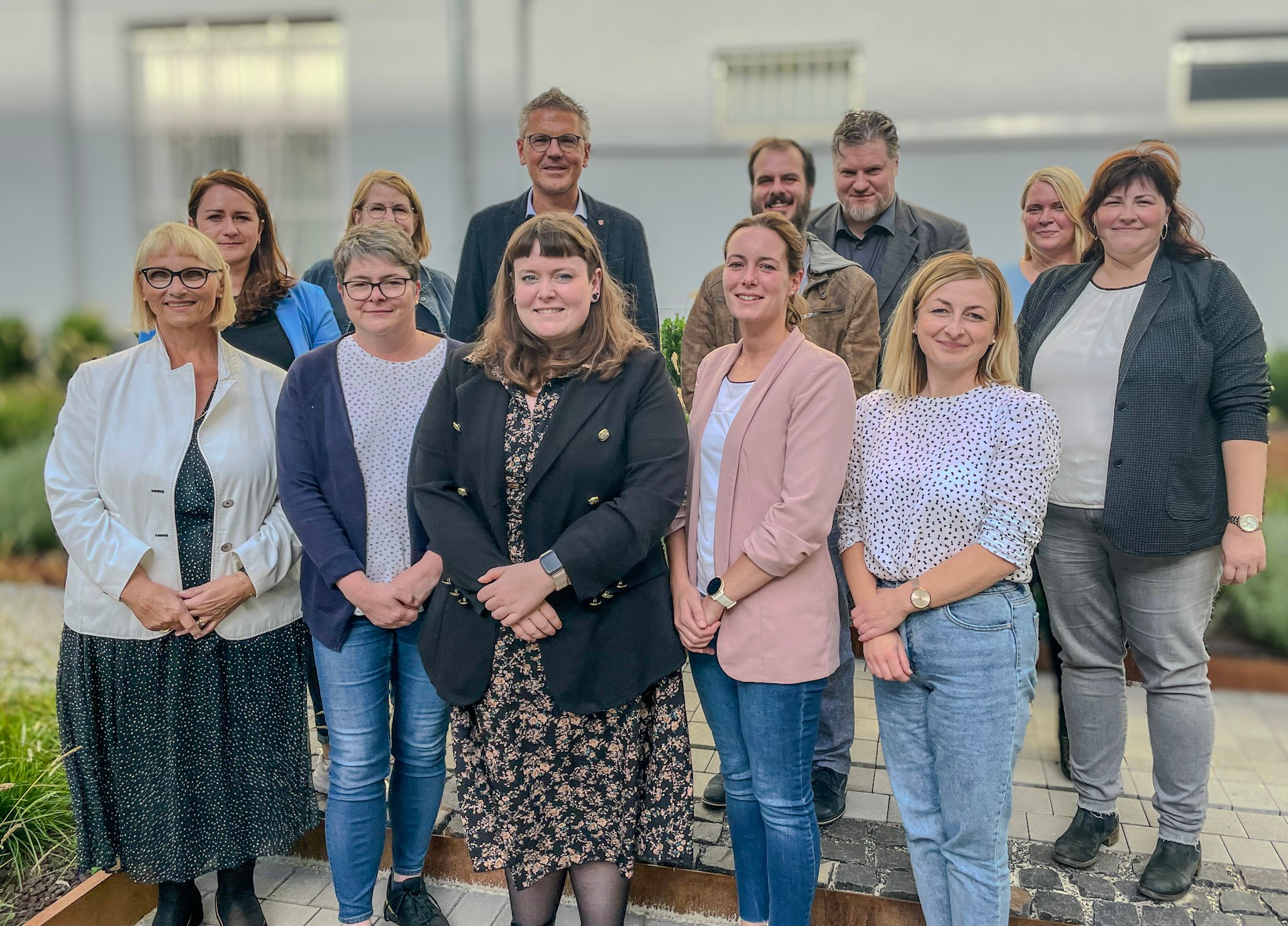 Auf dem Bild ist eine Gruppe von 12 Personen zu sehen, die sich für ein Foto aufstellen. Sie stehen vor einem Gebäude, das im Hintergrund unscharf ist. Die Gruppe besteht aus unterschiedlich gekleideten Männern und Frauen, einige tragen formelle Business-Kleidung, während andere in legerer Kleidung sind. Sie stehen in zwei Reihen, wobei die hintere Reihe auf einer erhöhten Stufe steht, um besser sichtbar zu sein. Die Personen lächeln und scheinen für ein gemeinschaftliches Event oder ein Teamfoto zusammengekommen zu sein. Die Stimmung wirkt freundlich und kooperativ.