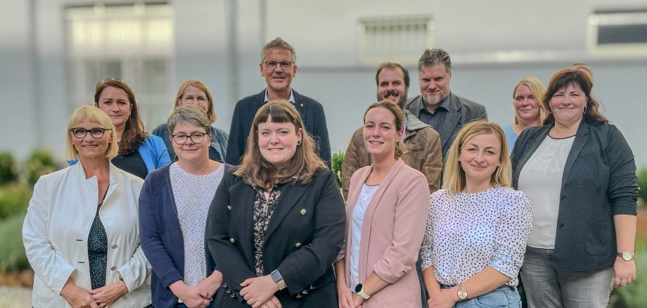 Auf dem Bild ist eine Gruppe von 12 Personen zu sehen, die sich für ein Foto aufstellen. Sie stehen vor einem Gebäude, das im Hintergrund unscharf ist. Die Gruppe besteht aus unterschiedlich gekleideten Männern und Frauen, einige tragen formelle Business-Kleidung, während andere in legerer Kleidung sind. Sie stehen in zwei Reihen, wobei die hintere Reihe auf einer erhöhten Stufe steht, um besser sichtbar zu sein. Die Personen lächeln und scheinen für ein gemeinschaftliches Event oder ein Teamfoto zusammengekommen zu sein. Die Stimmung wirkt freundlich und kooperativ.
