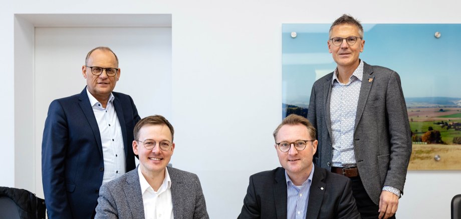 Zwei Männer sitzen an einem Tisch und halten Kugelschreiber in der Hand. Es sieht aus, als ob sie einen Vertrag unterzeichnen. Zwei Männer stehen dahinter. Alle lächeln in die Kamera. 