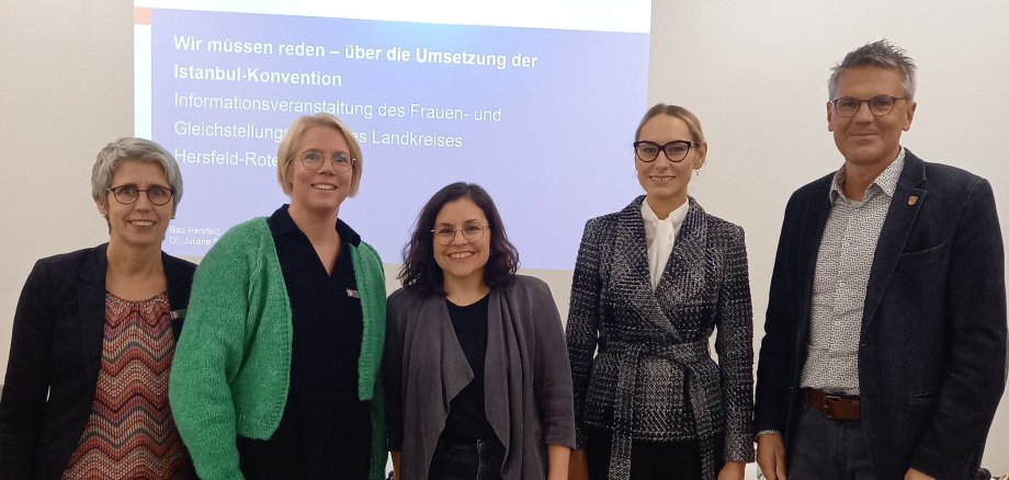 Vier Frauen und ein Mann stehen vor einer Wand auf der eine Power-Point-Präsentation gezeigt wird. Sie lächeln.