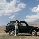 Eine Frau steht vor einem schwarzen Auto mitten in der bergigen Natur.