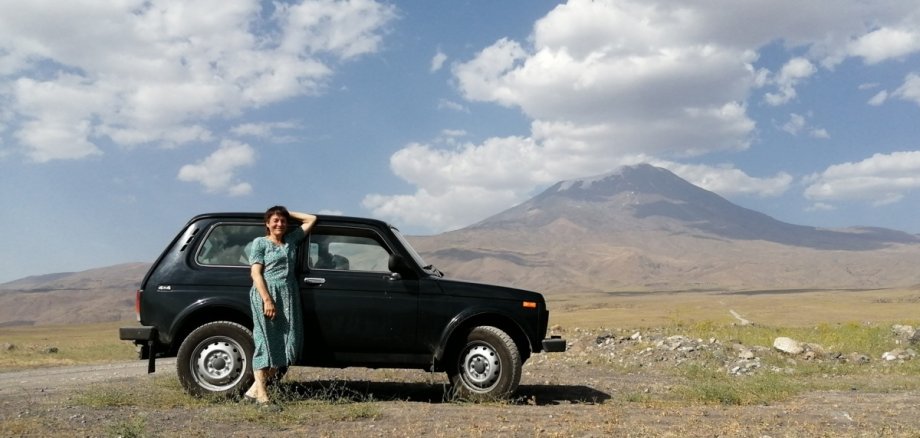 Eine Frau steht vor einem schwarzen Auto mitten in der bergigen Natur.
