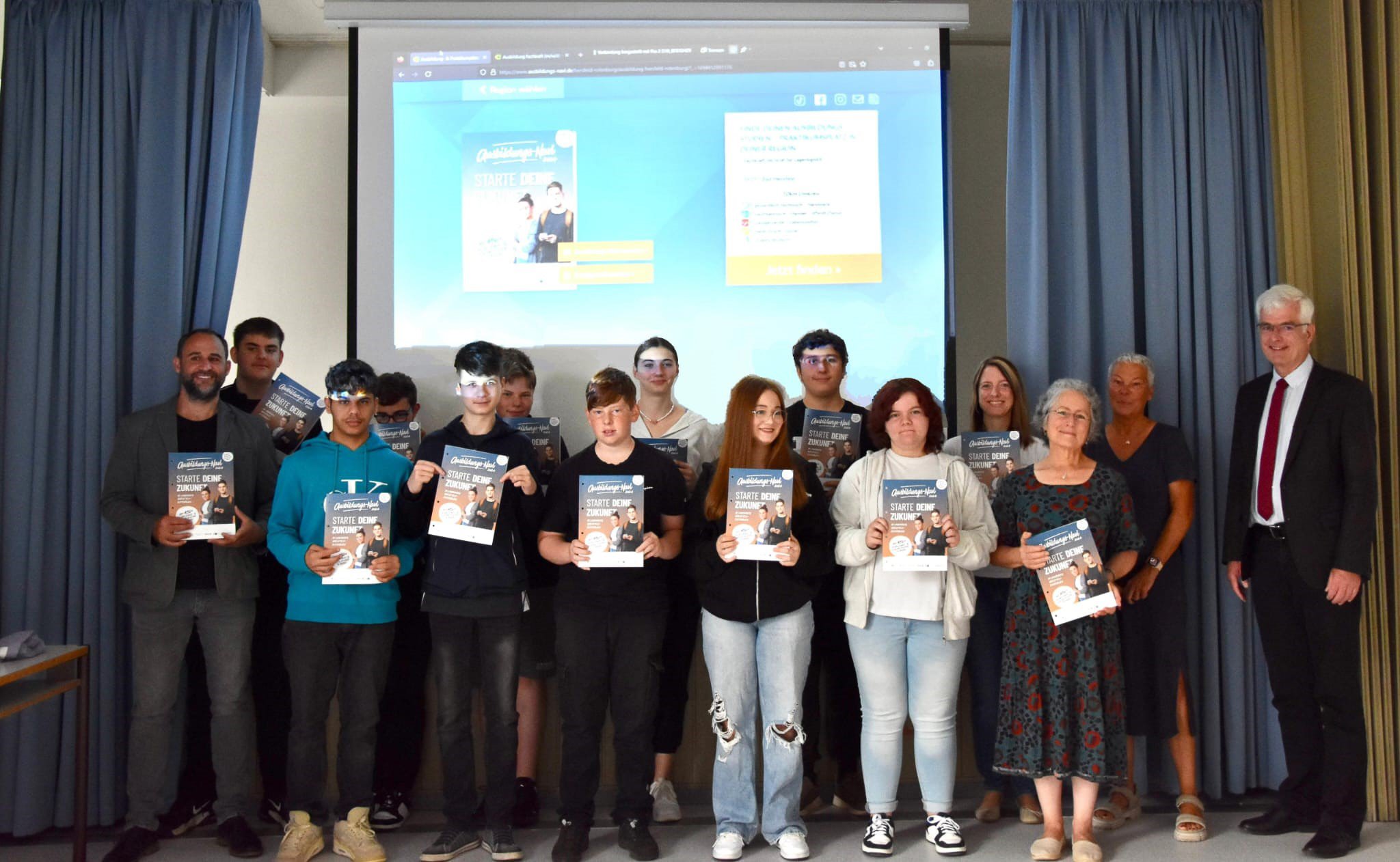 Gruppenfoto der Klasse Bo3 der Friedrich-Fröbel-Schule vor einer Leinwand mit Landrat Torsten Warnecke. Die Schülerinnen und Schüler halten das Ausbildungs-Navi in der Hand.