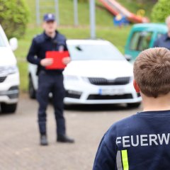 Ein Junge in einem Feuerwehr-T-Shirt steht im Vordergrund, während im Hintergrund ein Feuerwehrmann mit einem roten Klemmbrett zu sehen ist.