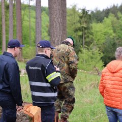 In einem Waldgebiet stehen mehrere Einsatzfahrzeuge der Feuerwehr. Ein Feuerwehrmann und ein Soldat schauen in die Ferne.