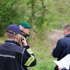 Eine Gruppe von Männern steht mit dem Rücken zum Betrachter im Wald, eine Person funkt.