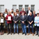 Ungefähr 15 Personen stehen im Innenhof der Kreisverwaltung. Einige davon halten Zertifikate in der Hand, die sie im Zuge der Fortbildung zum Vermittlungscoach erhalten haben. Alle Personen sind glücklich und lächeln.