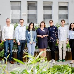 Ein Gruppenfoto von neun Personen, die in einem Garten stehen. Sie tragen Namensschilder und lächeln in die Kamera.