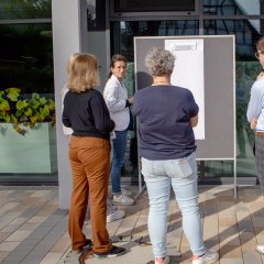 Mehrere Personen stehen um ein Whiteboard herum, das draußen aufgestellt ist. Sie diskutieren und einige notieren Punkte auf dem Board.