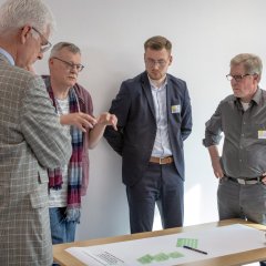 Eine Gruppe von Männern diskutiert um einen Tisch herum, wobei eine Person auf ein Dokument zeigt. Sie wirken konzentriert und tauschen Ideen in einer Workshop-Umgebung aus.