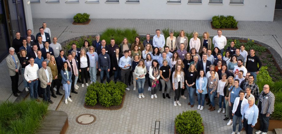 Eine große Gruppe von Personen steht für ein Gruppenfoto zusammen. Sie befinden sich in einem Innenhof eines modernen Gebäudes, das von Pflanzen und Bäumen umgeben ist.