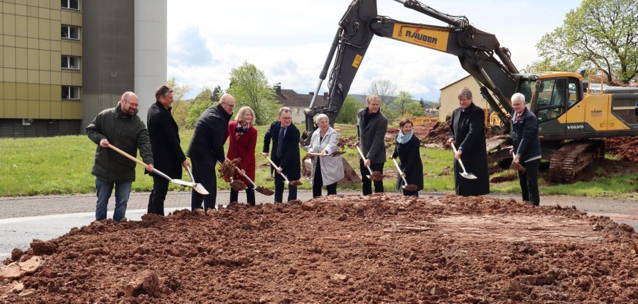 Auf dem Foto ist eine Gruppe von Personen zu sehen, die offenbar an einer zeremoniellen ersten Spatenstich-Zeremonie teilnehmen, was oft den Beginn von Bauprojekten markiert. Sie stehen nebeneinander auf einem Stück umgegrabenen Erdbodens, jeder hält eine Schaufel und scheint gerade eine Schaufel Erde zu heben oder zu bewegen. Im Hintergrund sind Baumaschinen zu sehen, darunter ein Bagger, der auf eine größere Baustelle hinweist. Die Umgebung deutet darauf hin, dass es sich um ein städtisches oder Vorstadtbereich handelt, und das Gebäude im Hintergrund könnte ein Teil des Klinikums sein, auf das im Bildtitel verwiesen wird. Die Personen sind formell gekleidet, was typisch für solche Anlässe ist, bei denen Beamte oder Würdenträger anwesend sein könnten.