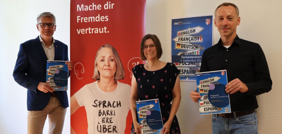 Zwei Männer und eine Frau stehen in einem Büro in der Kreisverwaltung. Es steht ein vhs-Banner zwischen zwei Personen und es hängt ein vhs-Plakat an der Wand. Die drei Personen lächeln in die Kamera und halten ein vhs-Programmheft in der Hand.