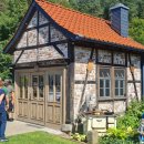 Auf dem Bild ist ein kleines Fachwerkhaus mit einem roten Ziegeldach zu sehen, das sich in einer ländlichen Umgebung befindet. Vor dem Haus stehen mehrere Personen, die sich das Gebäude ansehen. Das Haus hat große Holztüren und Fenster mit Sprossen. Vor dem Haus steht ein altes, cremefarbenes Küchenherd und ein kleines Gartenbeet mit bunten Blumen, in dem ein Gartenzwerg mit einer Schaufel steht. Im Hintergrund sind dichte, grüne Bäume und ein weiteres Gebäude mit einem ähnlichen Dach zu erkennen. Die Szene wirkt friedlich und idyllisch.