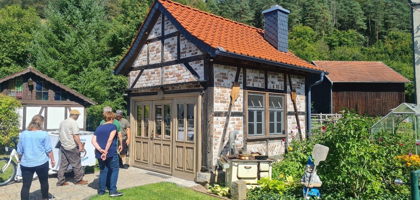 Auf dem Bild ist ein kleines Fachwerkhaus mit einem roten Ziegeldach zu sehen, das sich in einer ländlichen Umgebung befindet. Vor dem Haus stehen mehrere Personen, die sich das Gebäude ansehen. Das Haus hat große Holztüren und Fenster mit Sprossen. Vor dem Haus steht ein altes, cremefarbenes Küchenherd und ein kleines Gartenbeet mit bunten Blumen, in dem ein Gartenzwerg mit einer Schaufel steht. Im Hintergrund sind dichte, grüne Bäume und ein weiteres Gebäude mit einem ähnlichen Dach zu erkennen. Die Szene wirkt friedlich und idyllisch.