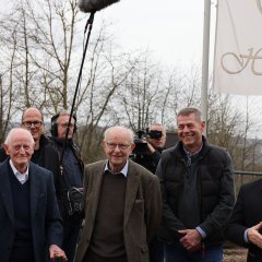 Eine Gruppe von Männern steht im Freien für ein Foto zusammen. Einige halten Kameras, während andere lächeln. Im Hintergrund weht eine Fahne mit der Aufschrift "Hotel".
