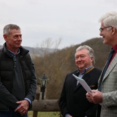 Drei Männer stehen im Freien und unterhalten sich. Der Mann in der Mitte lächelt, während der links im karierten Anzug steht und der rechts eine Kamera hält.