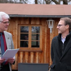 Zwei Männer in Winterkleidung unterhalten sich im Freien. Der eine trägt einen karierten Anzug und hält Dokumente, der andere trägt eine Jacke und eine Jeans.