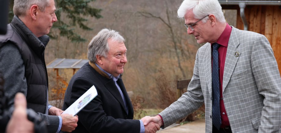 Zwei Männer geben sich die Hand. Der eine trägt einen karierten Anzug und eine rote Krawatte, der andere einen dunklen Mantel und ein grünes Dokument. Im Hintergrund ist eine Holzhütte zu sehen.