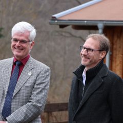Zwei Männer, einer im karierten Anzug und der andere in einem dunklen Mantel, stehen im Freien und unterhalten sich. Der Mann im Mantel trägt eine Brille.