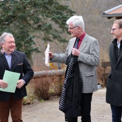 Zwei ältere Männer, einer mit einem Spazierstock und der andere mit einer Wurst in der Hand, stehen draußen. Zwei andere Männer schauen zu, einer lacht.