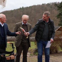Zwei ältere Männer, einer mit einem Spazierstock und der andere mit einer Wurst in der Hand, stehen draußen. Zwei andere Männer schauen zu, einer lacht.