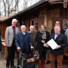 Eine Gruppe von sieben Personen steht vor einer Holzhütte. Zwei der Männer tragen Anzüge, einer davon ist mit einem Spazierstock ausgestattet. Eine Frau steht in der Mitte der Gruppe, sie trägt eine braune Jacke und hält eine Handtasche. Die Personen lächeln in die Kamera.