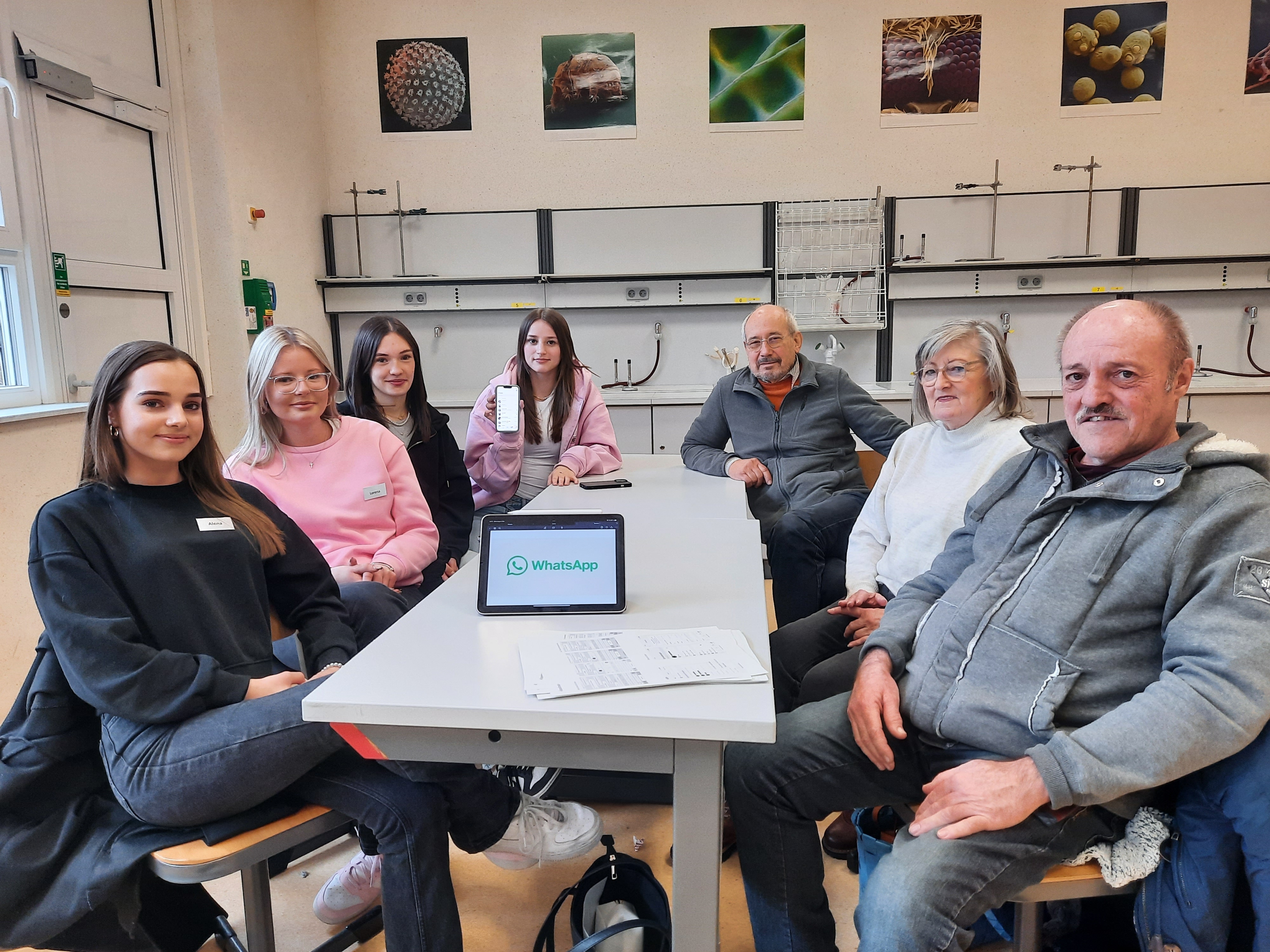 Vier Schülerinnen der Gesamtschule Niederaula und drei Senioren sitzen gemeinsam an einem Tisch in einem Unterrichtsraum. Auf dem Tisch steht ein Tablet mit der Aufschrift WhatsApp, eine Schülerin hält ein Handy in der Hand.