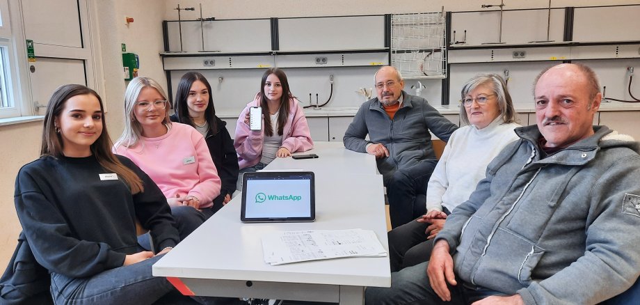 Vier Schülerinnen der Gesamtschule Niederaula und drei Senioren sitzen gemeinsam an einem Tisch in einem Unterrichtsraum. Auf dem Tisch steht ein Tablet mit der Aufschrift WhatsApp, eine Schülerin hält ein Handy in der Hand.