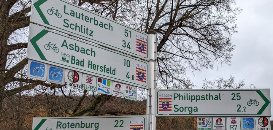 Ein Fahrradwegweiser im Landkreis Hersfeld-Rotenburg. Er zeigt an, wie weit die Distanz zu verschiedenen Orten im Landkreis ist. Im Hintergrund ist ein Baum ohne Blätter zu sehen. Es ist Winter.