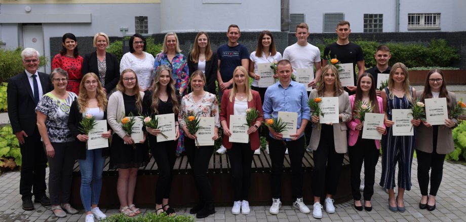 Die Auszubildenden, Verwaltungsfachwirte und Studierenden der Kreisverwaltung stehen im Innenhof der Kreisverwaltung. Sie halten Zertifikate und Blumen in der Hand.