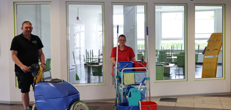 Ein Mann und eine Frau stehen in einem Schulflur vor der Cafeteria. Der Mann, der links steht, ist dunkel gekleidet und steht an einem Kehrwagen. Die Frau, die ein rotes T-Shirt an hat, steht hinter einem gut ausgestatteten Putzwagen.
