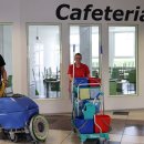 Ein Mann und eine Frau stehen in einem Schulflur vor der Cafeteria. Der Mann, der links steht, ist dunkel gekleidet und steht an einem Kehrwagen. Die Frau, die ein rotes T-Shirt an hat, steht hinter einem gut ausgestatteten Putzwagen.