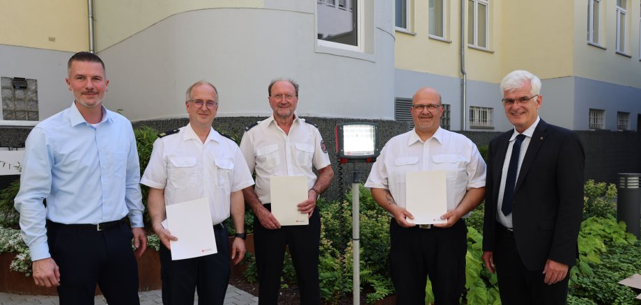 Die fünf Männer stehen draußen vor einem Gebäude und posieren für ein Foto. Drei von ihnen tragen weiße Uniformhemden und schwarze Hosen, während zwei Männer, die links und rechts außen stehen, formelle Kleidung tragen – der linke Mann trägt ein hellblaues Hemd und der rechte Mann einen schwarzen Anzug. Die drei Männer in der Mitte halten jeweils ein Dokument in der Hand. Im Hintergrund sind Pflanzen und ein mehrstöckiges Gebäude mit mehreren Fenstern zu sehen. Eine mobile Leuchte auf einem Stativ steht in der Mitte der Gruppe.