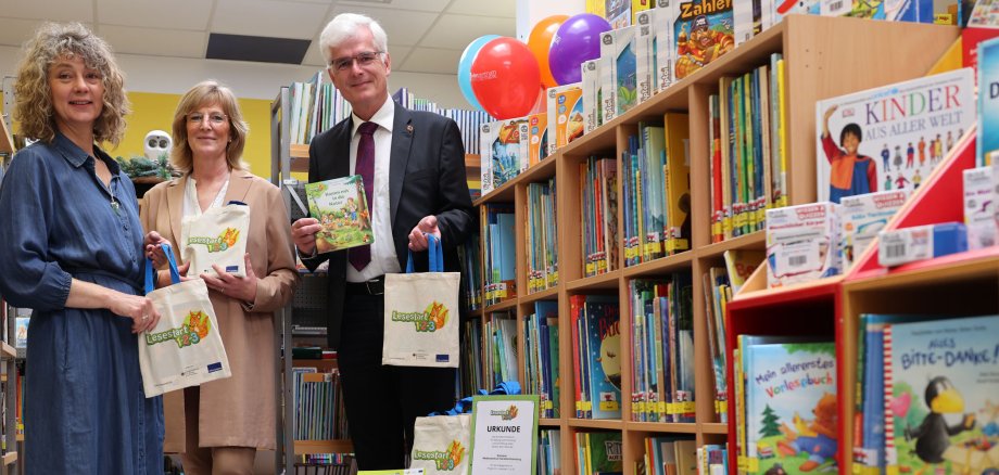 Drei Personen stehen im Medienzentrum des Landkreises. Am rechten Bildrand sieht man ein großes Regal, das mit vielen Büchern gefüllt ist. Die Personen halten kleine Taschen in der Hand.