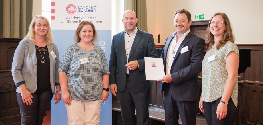 Gruppenfoto von fünf Personen, die in einem Innenraum vor einem Banner stehen. Auf dem Banner steht 'Land hat Zukunft - Akademie für den ländlichen Raum Hessen'. Eine Person hält eine Urkunde in der Hand, während die anderen lächeln. Alle tragen Namensschilder