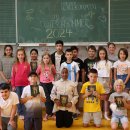 Rund 25 Schülerinnen und Schüler stehen vor einer Tafel in einem Klassenraum. Auf der Tafel steht mit Kreide geschrieben: "Herzlich willkommen im Deutschsommer 2024". Die meisten Kinder halten eine Ausgabe des Buches Ronja Räubertochter in der Hand.