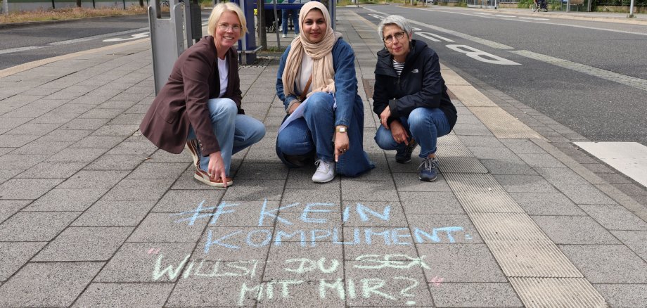 Die beiden Frauen- und Gleichstellungsbeauftragten und ihre Praktikantin hocken am Busbahnhof in Bad Hersfeld und zeigen auf einen Spruch, der mit Kreide auf den Boden gezeichnet wurde. Dort steht: "#KeinKompliment: Ich will Sex mit dir".