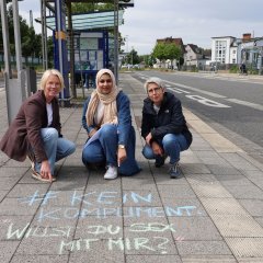 Die beiden Frauen- und Gleichstellungsbeauftragten und ihre Praktikantin hocken am Busbahnhof in Bad Hersfeld und zeigen auf einen Spruch, der mit Kreide auf den Boden gezeichnet wurde. Dort steht: "#KeinKompliment: Ich will Sex mit dir".