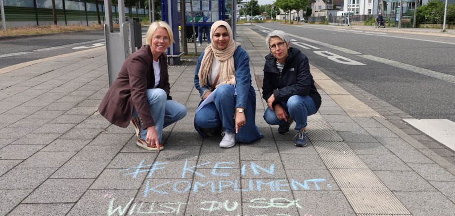 Die beiden Frauen- und Gleichstellungsbeauftragten und ihre Praktikantin hocken am Busbahnhof in Bad Hersfeld und zeigen auf einen Spruch, der mit Kreide auf den Boden gezeichnet wurde. Dort steht: "#KeinKompliment: Ich will Sex mit dir".