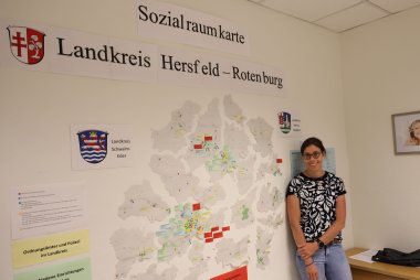 Eine Frau steht vor einer Wand in einem Büro im Landratsamt. Auf der Wand ist eine Sozialraum-Karte des Landkreises abgebildet. Die Frau lächelt in die Kamera.