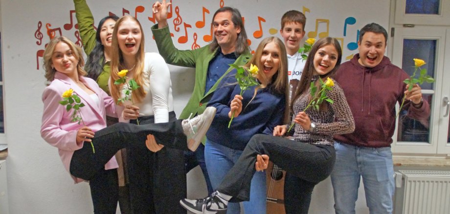 Ein Gruppenfoto von sieben fröhlichen Personen in einem Zimmer, die jeweils eine gelbe Blume halten und verschiedene freudige Posen einnehmen. Im Hintergrund sieht man Musiknoten an der Wand, was darauf hindeuten könnte, dass die Gruppe musikalisch aktiv ist oder eine Feier im Zusammenhang mit Musik stattfindet.