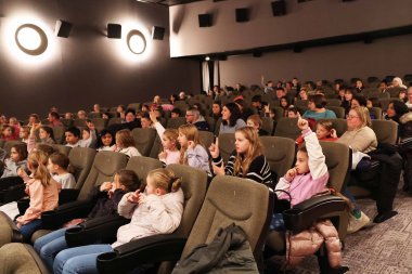 Viele Kinder und Erwachsene sitzen in einem Kino-Saal. Einige von ihnen heben die Hände.