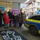 Einige Menschen stehen an der abgesperrten Tatort-Nachbildung. Auf der rechten Seite sieht man ein Polizei-Auto. 