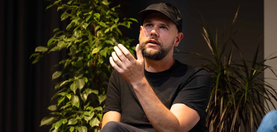 Ein Mann mit einer schwarzen Cap und einem schwarzen T-Shirt sitzt auf einem Stuhl.