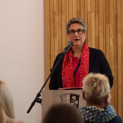 Eine Frau mit einem pinken Schal steht hinter einem Mikrofon und sagt etwas.