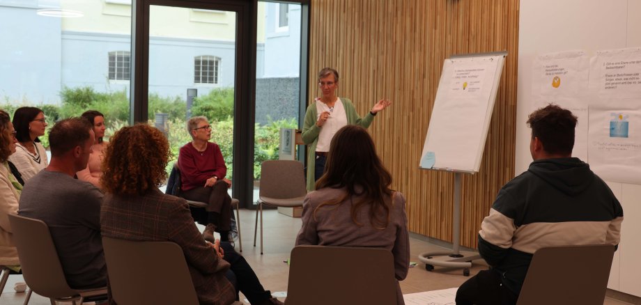 Mehrere Leute sitzen in einer Sitzgruppe im Kreistagssitzungssaal des Landkreises Hersfeld-Rotenburg. In der Nähe einer Flipchart steht eine Frau.