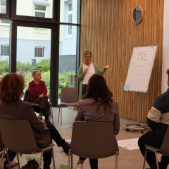 Mehrere Leute sitzen in einer Sitzgruppe im Kreistagssitzungssaal des Landkreises Hersfeld-Rotenburg. In der Nähe einer Flipchart steht eine Frau.